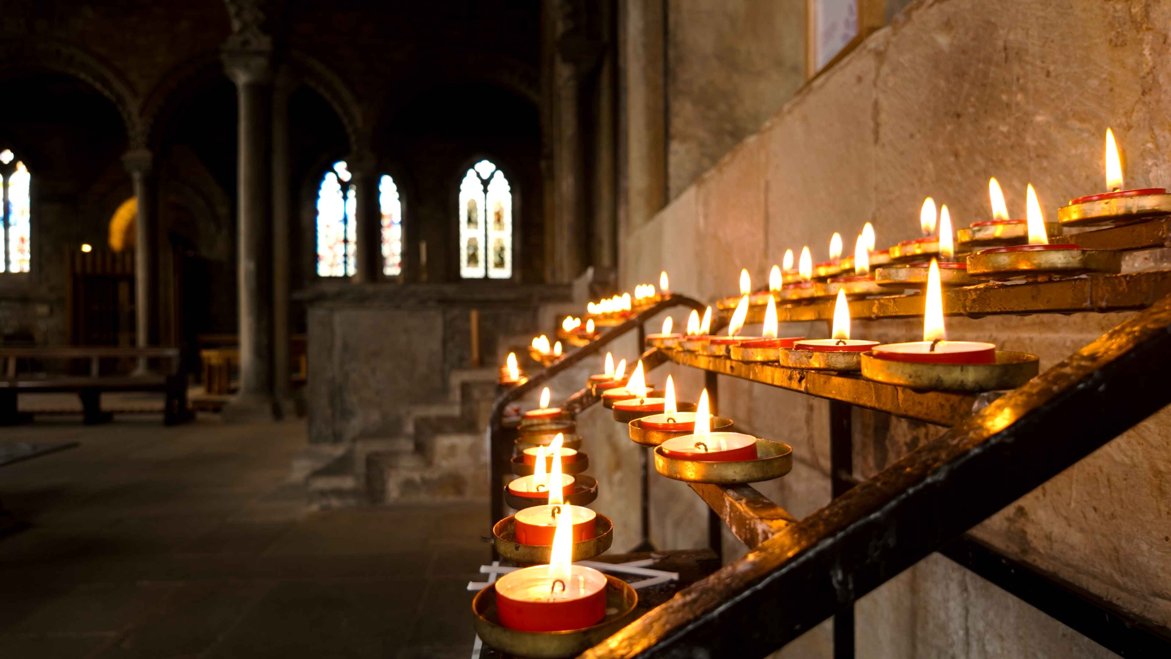 Several lit prayer candles on a stand with stained glass windows in the backdrop