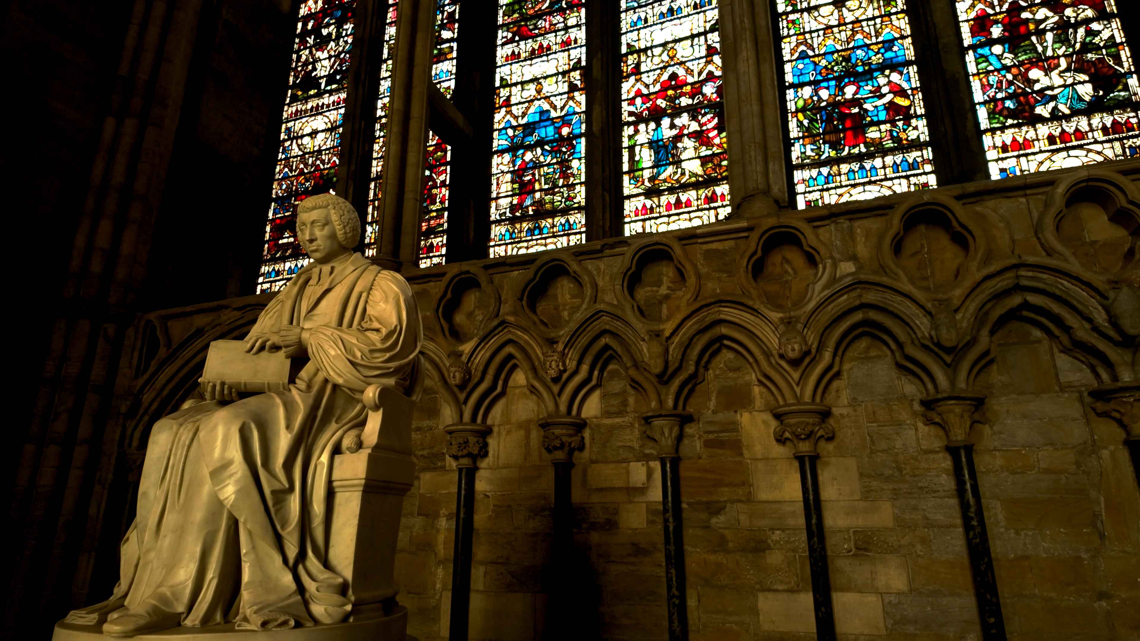 A statue of a man holding a book seated in front of a series of colorful stained glass window
