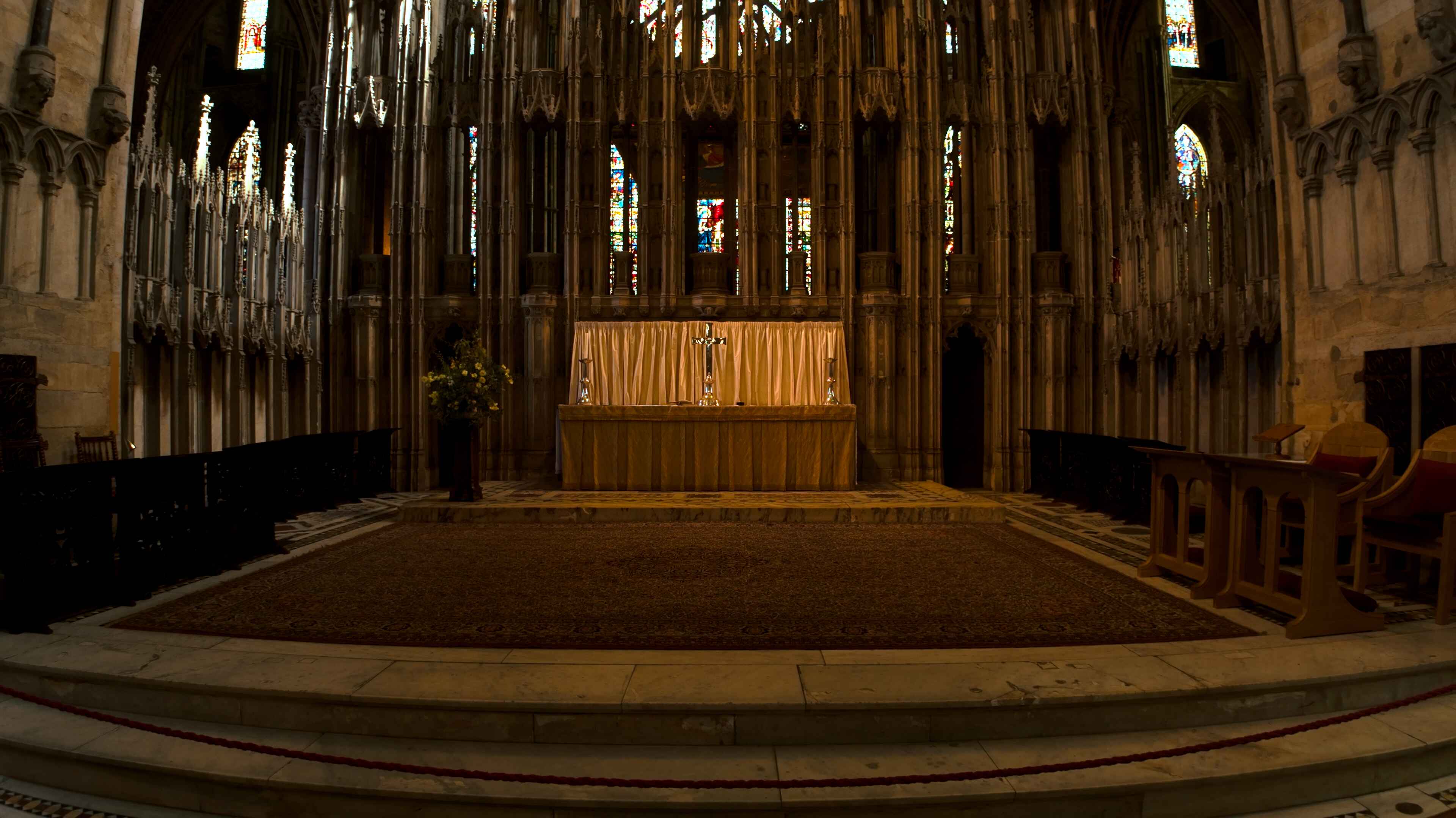 A church alter with the cross on a stand and large stained glass windows in the backdrop