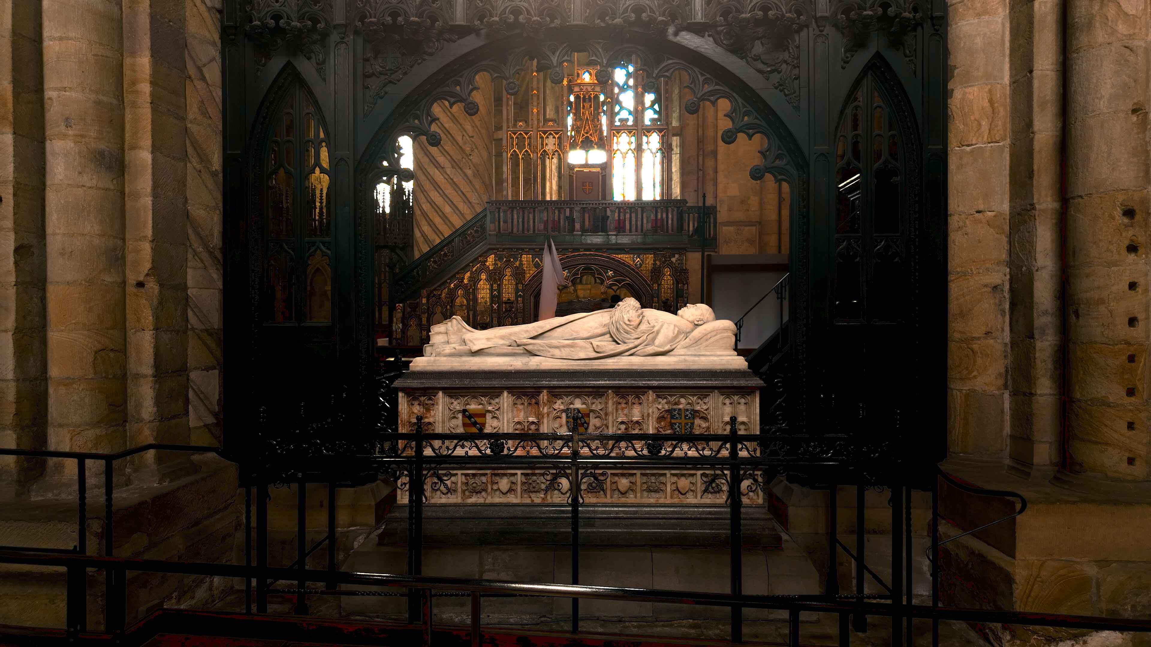 A stone tomb with reclining effigies is displayed inside a cathedral, surrounded by ornate ironwork and stained glass windows in the background